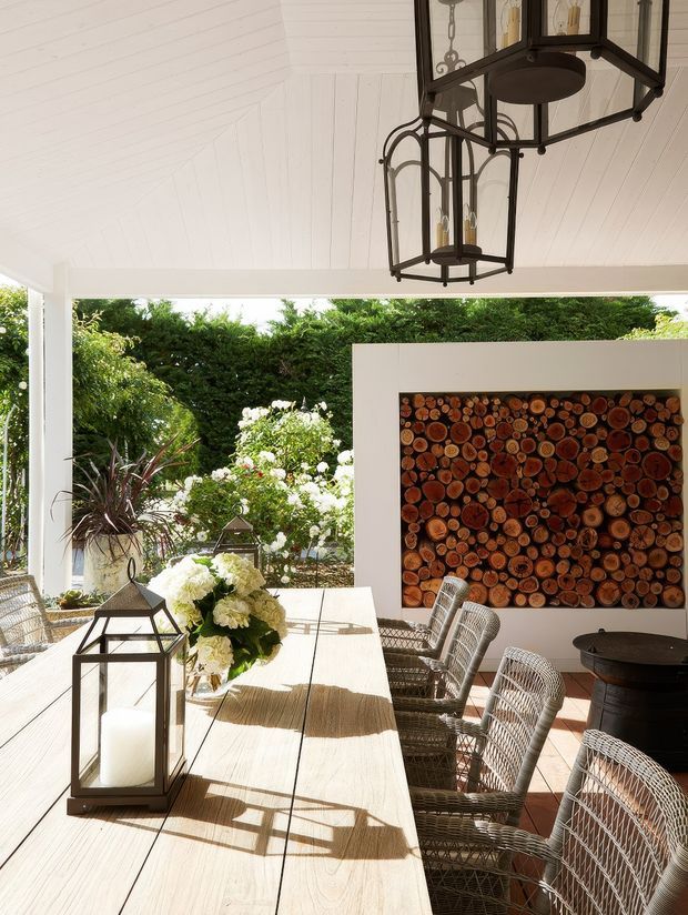 Bright covered outdoor dining area with a long pale timber dining table, woven wicker dining chairs, black metal lanterns, and a large decorative wall installation made from cut wood rounds. The space opens onto a lush garden and is sheltered by a white timber-lined ceiling with exposed dark metal pendant lanterns. The overall feel is relaxed, natural, and refined.