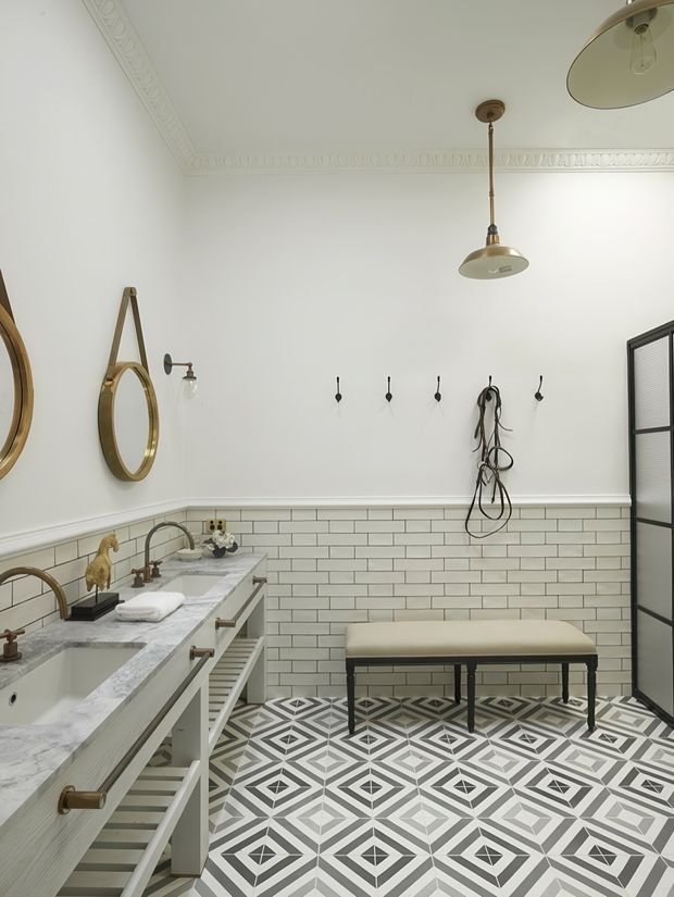 A refined bathroom with a long marble-topped vanity, twin basins, brass tapware, white subway-tile wainscoting, ornate ceiling cornices, geometric patterned floor tiles, round hanging mirrors, wall hooks, and a freestanding upholstered bench. The palette is predominantly white, light grey, and brass with a classic European-inspired character.