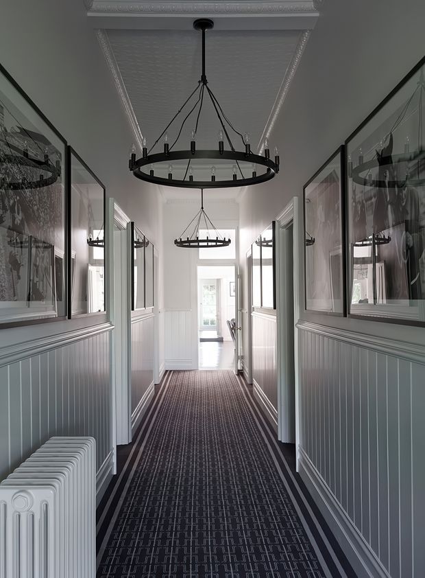 A long, narrow traditional hallway with dark patterned carpet, grey-blue beadboard wainscoting, white trim and ceiling mouldings, framed artwork, multiple black circular pendant chandeliers, and a white cast-iron radiator. Glazed doorways and bright windows lead toward the far end of the corridor.