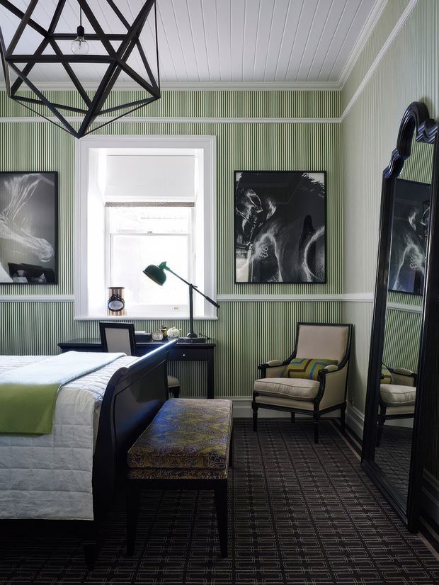 A refined bedroom with green vertically striped wallcovering, white paneled ceiling and detailed trim, dark patterned carpet, and a large black-framed mirror. The room contains a dark wood bed with white bedding and a pale green throw, a patterned upholstered bench, a classic armchair, a compact desk with task lamp, framed monochrome artwork, and a geometric black pendant light. The overall composition is richly layered, tailored, and traditional with contemporary decorative elements.