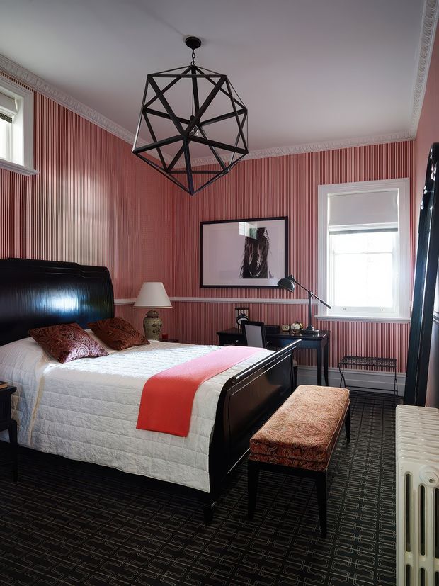 A richly styled bedroom with red-and-white vertically striped wallpaper, ornate ceiling cornice, dark patterned carpet, and a large black geometric pendant light. The room features a dark wood or black sleigh-style bed with white quilted bedding, red patterned cushions and throw, a matching upholstered bench, framed artwork, a small desk, windows with blinds, and a traditional radiator.