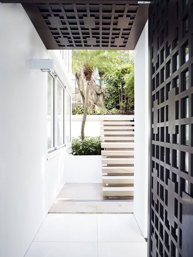 A bright, narrow architectural passage leading to a floating staircase and small planted courtyard. White walls and large pale floor tiles contrast with dark geometric breeze-block screens at the entrance and overhead, creating a strong patterned architectural feature. Tropical planting is visible beyond the stairs.