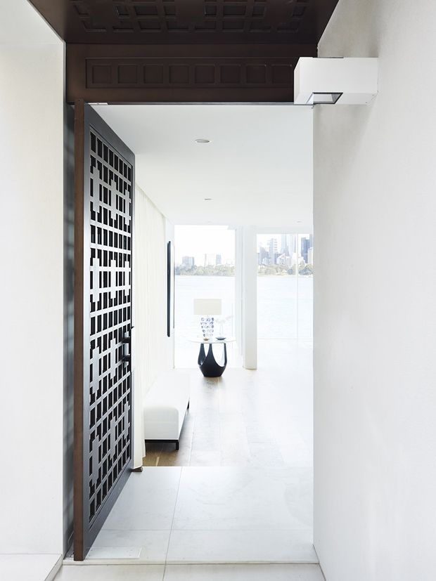 A bright, minimalist interior view through a hallway or entry passage into a waterfront-facing living space. The foreground features a dramatic dark timber ceiling with recessed square detailing and a matching geometric timber screen door or partition. White walls, pale tiled flooring, recessed ceiling lights, and a sculptural black side table create a high-contrast contemporary setting. Large floor-to-ceiling glazing at the far end overlooks water and a city skyline.