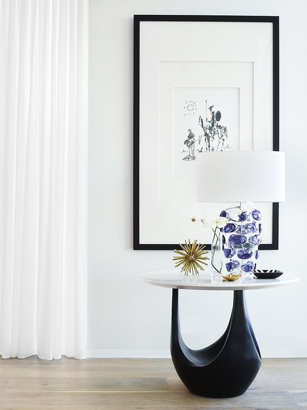 A bright, refined interior vignette featuring a slim white console table with a sculptural black pedestal base, styled with a modern lamp, blue-and-white vase, metallic starburst ornament and small bowl. A large black-framed monochrome artwork hangs on the white wall behind it, with sheer white curtains at left and pale timber flooring below.
