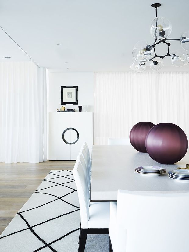 Bright, contemporary dining room with a long white dining table and white upholstered chairs, set over a light wood floor and geometric black-and-white rug. Sheer full-height curtains cover the windows, while a sculptural glass globe chandelier and deep purple spherical decorative objects provide focal points. A white sideboard with a circular black detail sits against the rear wall.