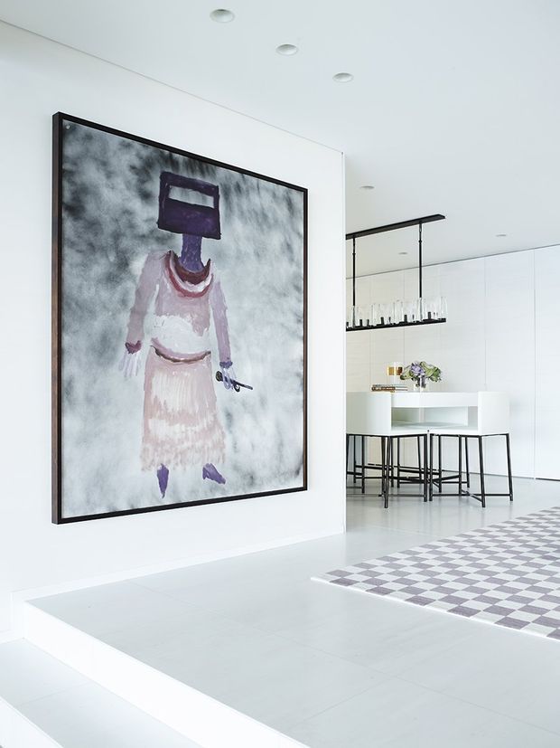 Bright, minimalist open-plan interior with a white tiled or stone floor, white walls and ceiling, and recessed downlights. A large framed abstract artwork dominates the foreground. Beyond the partial dividing wall is a contemporary kitchen or dining area with white full-height cabinetry, a white island with open shelving, black metal bar stools, and a linear black pendant light. A geometric grey-and-white checkered rug adds contrast.