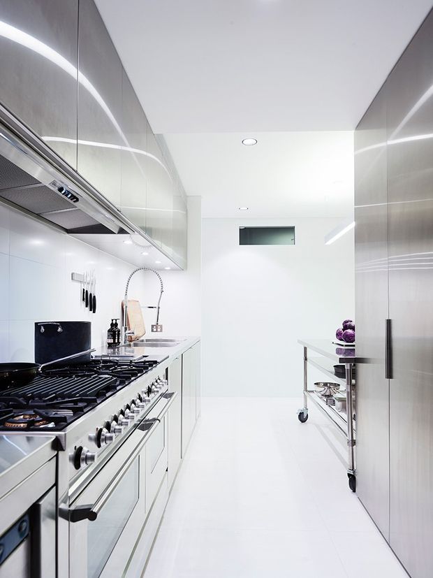 Bright, narrow contemporary galley kitchen with extensive stainless-steel appliances and cabinetry, white tiled walls and flooring, glossy reflective surfaces, a large professional range, and a movable stainless-steel prep cart.