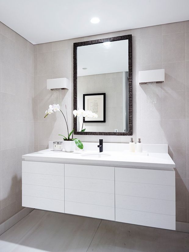 A contemporary bathroom vanity area with a wide floating white double-sink cabinet, white benchtop, wall-mounted black tapware, pale large-format tiled walls and floor, and a large dark textured-framed mirror. Minimal white wall sconces flank the mirror, with orchids and small bathroom accessories on the vanity.