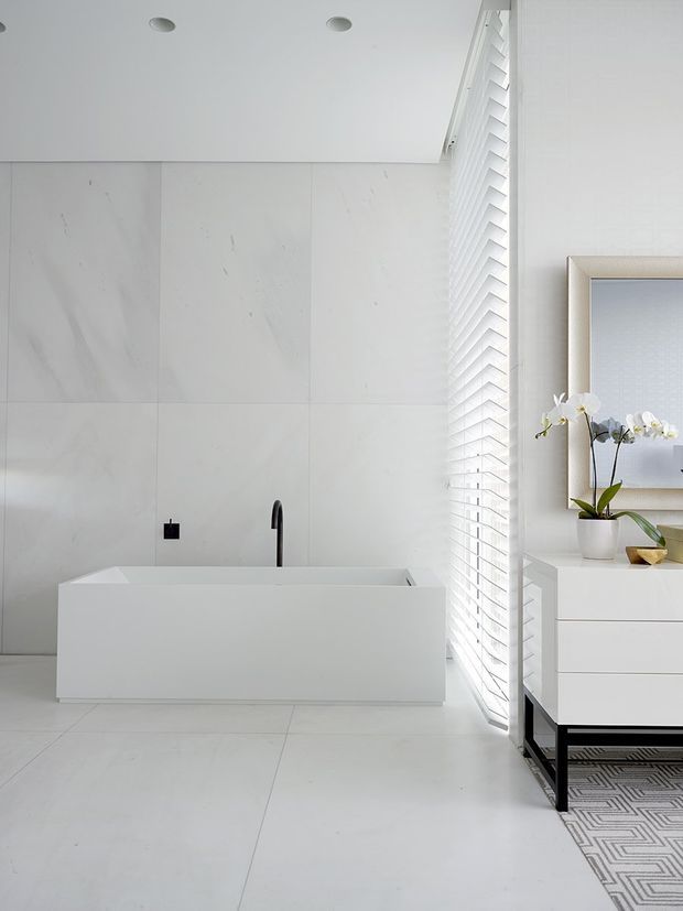 Bright, minimalist white bathroom featuring a freestanding rectangular bathtub with a matte black wall-mounted faucet, large white marble-look wall and floor tiles, and white louvred window blinds. A modern white vanity with black base and countertop styling is visible on the right, along with an orchid and framed artwork. The room has abundant natural light and a clean, serene atmosphere.