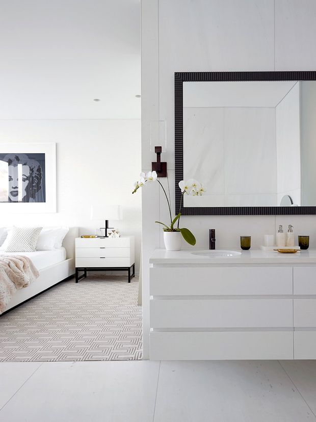 Bright, refined bathroom vanity photographed beside an adjacent bedroom. The bathroom features a wall-mounted white vanity with integrated basin, chrome fittings, a large dark ribbed-framed mirror, and a white orchid. The adjoining bedroom has a white upholstered bed, white bedside table, monochrome artwork, and a geometric patterned carpet. The overall scheme is predominantly white with restrained black and grey accents.