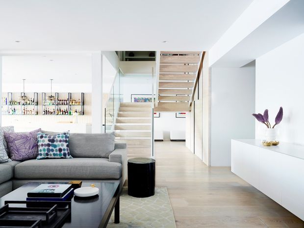 Bright open-plan contemporary living space with a grey sectional sofa and black coffee table in the foreground, a minimalist white built-in console, pale timber flooring, and a floating open-tread staircase leading upward. A bar or display area with bottles is visible in the background, while purple cushions and flowers provide restrained colour accents.