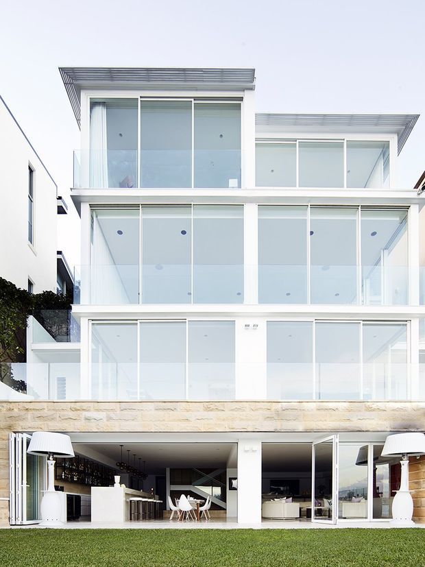 A bright multi-storey contemporary residential building viewed from the lawn, with extensive floor-to-ceiling glazing, white rendered walls, transparent glass balconies, projecting flat roof canopies, and a light stone retaining wall. The open lower level reveals a bar and living/dining area, while large white outdoor lamps flank the facade.
