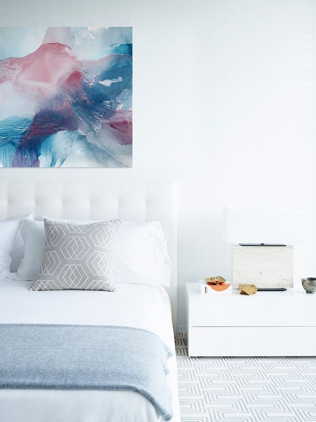 Bright, highly styled bedroom with a white upholstered tufted headboard, crisp white bedding, and a pale blue-grey throw. A geometric grey accent cushion echoes the patterned grey-and-white rug. The room includes a low white bedside cabinet with a pale stone or terrazzo-like lamp and small decorative objects, while a colourful abstract artwork in pink, blue, and white hangs above the bed. The overall look is clean, airy, cool-toned, and contemporary.