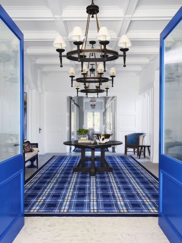 A bright, formal entry hall viewed through open vivid-blue double doors. The white paneled walls and coffered ceiling are lined with a dramatic series of dark metal chandeliers, while a dark round console table sits centrally on a large blue plaid rug. Blue woven chairs and decorative accessories add a refined, high-contrast decorative scheme.