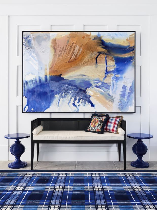 A refined interior vignette featuring a black-framed abstract artwork above a slim black bench with a cream upholstered seat and patterned cushions. Two matching deep-blue pedestal side tables flank the bench, set against white paneled walls. A bold blue plaid rug grounds the composition, creating a polished, graphic, layered look.