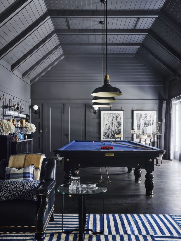 A dramatic, dark-toned billiards room with a traditional pool table beneath a large layered pendant light. The room features charcoal-painted timber wall and ceiling panelling, exposed truss-like ceiling framing, paneled double doors, framed artwork, a built-in drinks display, a black leather sofa with patterned cushions, and a blue-and-white striped rug. Natural light enters through tall glazing on the right.