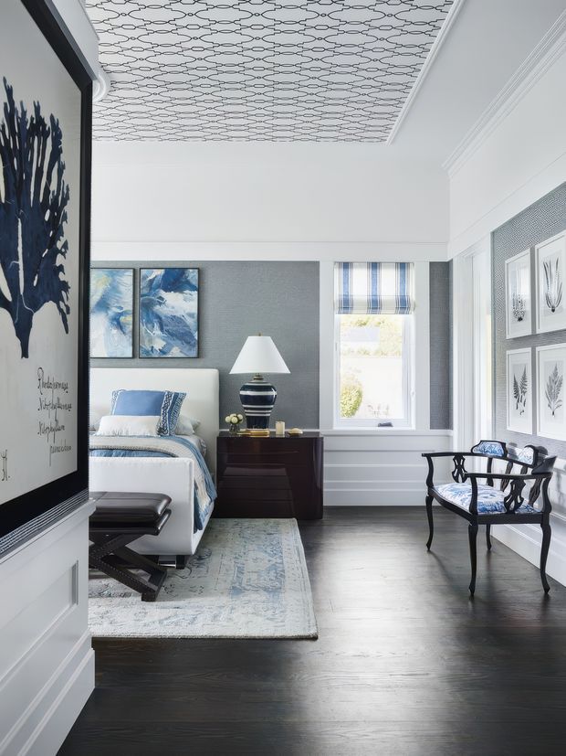 A refined bedroom with a white upholstered bed, blue-and-white bedding, dark timber flooring, and a dark wood bedside cabinet. The room features a grey-blue textured accent wall, patterned wallpapered ceiling, white decorative wall panelling and moulded cornices, framed botanical artwork, a striped Roman blind, and an elegant dark-framed armchair. The palette is predominantly white, grey, blue, and dark wood, creating a polished traditional coastal feel.