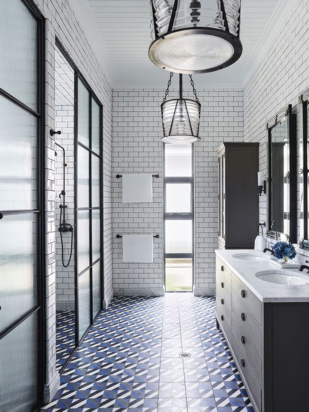 Bright, narrow contemporary bathroom with white subway-tile walls, a geometric blue, white and grey patterned tile floor, black-framed glass shower enclosure, double vanity with pale cabinetry and white stone countertop, tall window, mirrored panels, and distinctive industrial-style pendant lights.