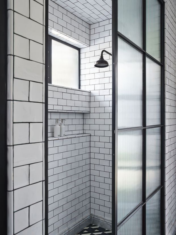 A compact walk-in shower with white rectangular subway tiles covering the walls and ceiling, dark grout, black metal framing, and a black rainfall showerhead. A translucent ribbed-glass partition and door define the shower enclosure, with a narrow built-in tiled shelf holding toiletries. The space has a clean, monochromatic industrial-modern aesthetic.