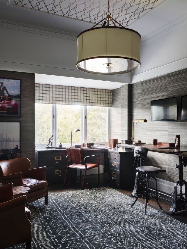 A sophisticated home office with a dark built-in desk wrapping beneath a large window, black filing drawers, leather desk chair and armchairs, wall-mounted screens, and a bar-height work surface. The room features dark textured wallcovering, white architectural trim, a patterned ceiling, a large geometric area rug, and a prominent drum pendant light. Natural daylight creates a refined, moody atmosphere.