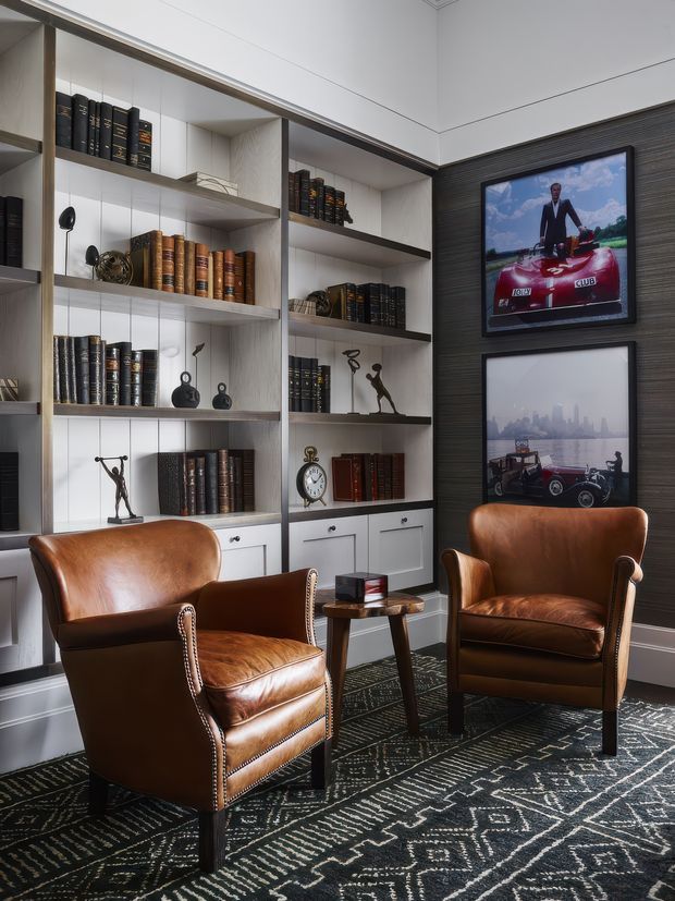 A refined study or library nook with floor-to-ceiling built-in shelving filled with books and decorative objects, two cognac leather armchairs, a small wooden side table, framed artwork, and a dark geometric patterned rug. The cabinetry combines white built-in storage with a dark wood-look feature wall, creating a sophisticated traditional-contemporary reading space.