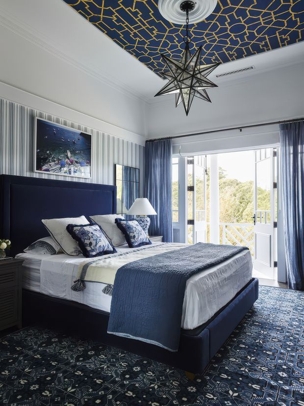 A richly decorated bedroom with a navy upholstered bed, layered white and blue bedding, patterned blue-and-white pillows, and a large patterned area rug. The walls are white with vertical panel detailing, while the ceiling features a bold navy geometric pattern with gold lines. A dramatic star-shaped pendant light hangs centrally, and full-height blue curtains frame glazed French doors opening to a bright exterior balcony or terrace. The overall look is luxurious, traditional, and highly patterned.