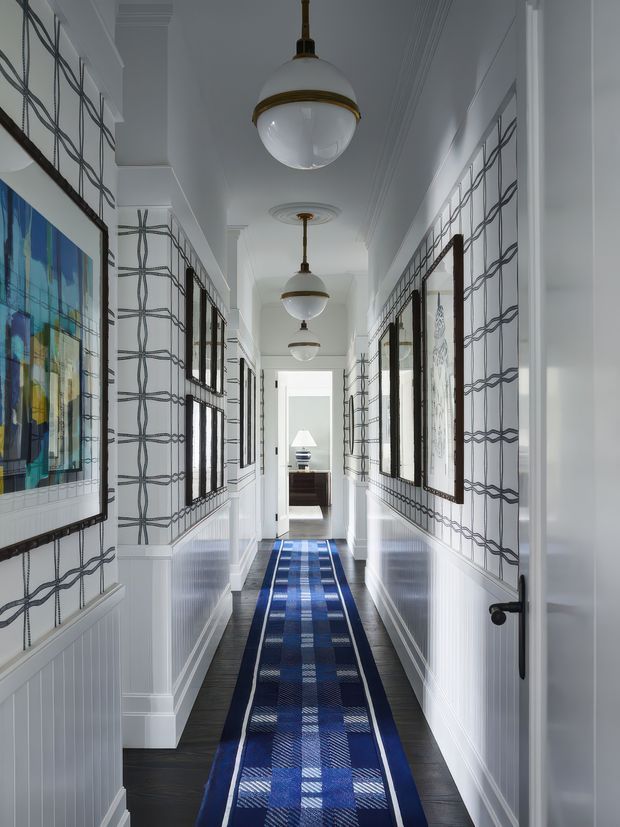 Long, narrow, bright residential hallway with white wainscoted walls and patterned wallpaper, dark timber flooring, multiple framed artworks and mirrors, and a blue patterned runner. Repeated pendant lights with brass detailing hang along the corridor, while decorative ceiling mouldings and multiple doorways create a refined, highly symmetrical composition.
