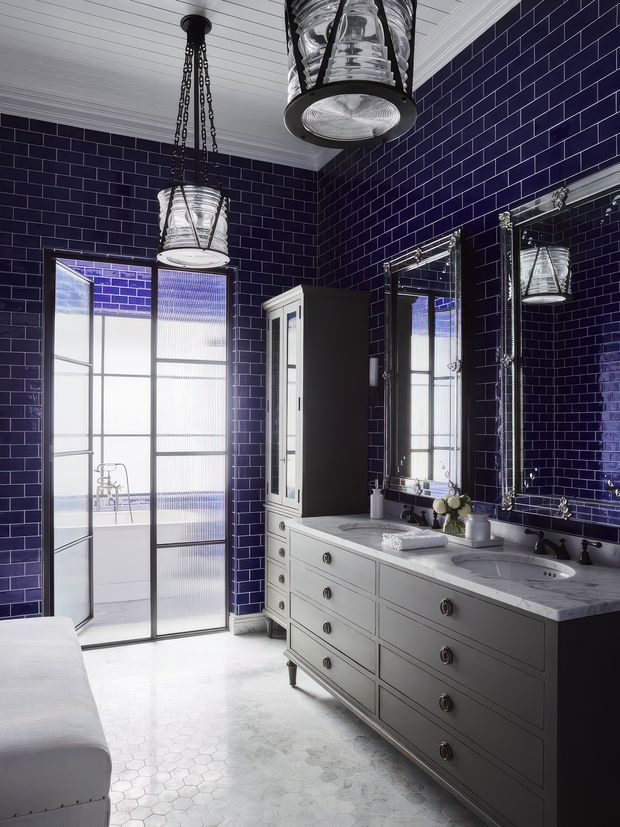 A sophisticated bathroom with deep navy-blue glossy subway-tiled walls, pale hexagonal floor tiles, a long grey-white double vanity with marble-like benchtop and twin basins, ornate framed mirrors, a tall storage cabinet, industrial nautical-style pendant lights, and a frosted glass door or shower enclosure. The room has a dramatic, refined monochromatic palette with metallic accents.