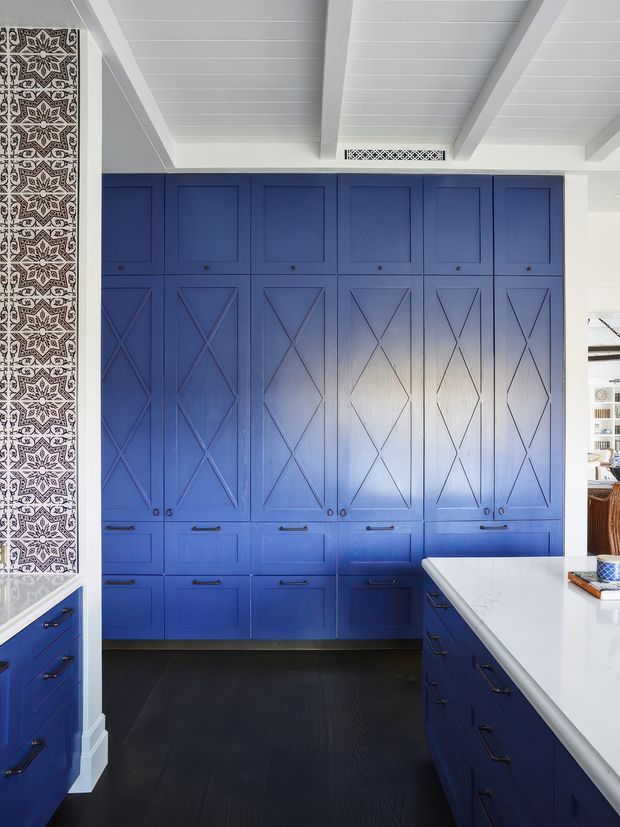 A bright, highly detailed kitchen featuring extensive deep blue built-in cabinetry with traditional framed panel doors and decorative X detailing. A white island with blue drawer cabinetry occupies the foreground, while patterned black-and-white tile forms a vertical feature beside the cabinetry. The room has a white painted timber-lined ceiling with exposed beams, dark flooring, and a light, refined overall scheme.