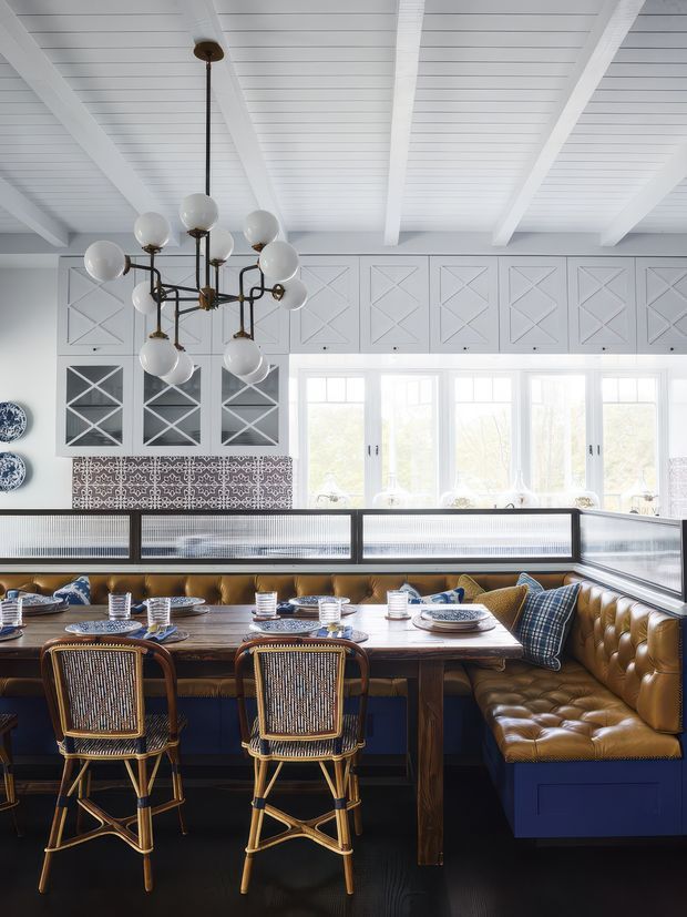 A bright, refined dining area with a built-in booth surrounding a substantial timber dining table. The booth has deep navy bases, tufted caramel leather upholstery, and patterned cushions, paired with woven rattan bar stools. White-painted cabinetry with geometric panel detailing, patterned tile backsplash, large windows, and a white-beamed plank ceiling create a layered classic coastal interior. A sculptural brass and glass globe chandelier hangs above the table.