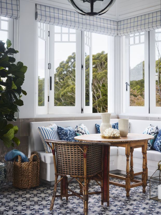 Bright, elegantly styled bay-window sunroom with white framed glazing, blue-and-white checked Roman blinds, a built-in upholstered window seat, patterned cushions, a rustic wooden dining table, woven cane chairs, decorative vessels, indoor plants, and a patterned tiled floor. The room has a fresh coastal and traditional character with abundant natural light and garden views.