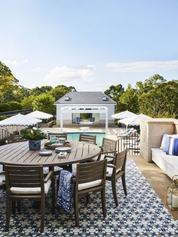 Bright, elevated outdoor entertaining terrace overlooking a rectangular swimming pool and a pavilion. The terrace features a large round timber dining table with matching chairs, patterned blue-and-white floor tiles, white umbrellas, a built-in pale masonry bench with blue-and-white cushions, black metal balustrades, and surrounding mature greenery. The pavilion has a dark pitched tiled roof and white columns, creating a resort-like outdoor setting.