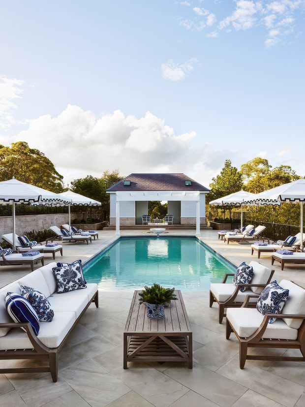 A symmetrical outdoor pool area with a long rectangular turquoise swimming pool, pale stone paving, and numerous white-cushioned wooden lounge chairs arranged around the edges. White parasols with dark trim provide shade, while a small pavilion with a tiled pitched roof and open seating area anchors the far end. The setting is bright, refined, and resort-like, with mature trees and blue sky in the background.