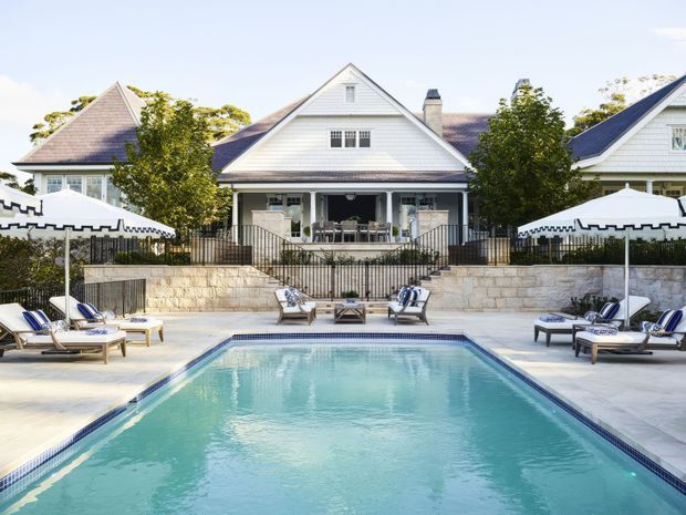 A symmetrical outdoor swimming pool courtyard in front of a large white weatherboard house. The pool is surrounded by pale stone paving, sun loungers, striped cushions, and large white umbrellas. Behind it, the multi-gabled residence features white timber cladding, dark roof shingles, covered verandas, black railings, and stone retaining walls. The scene is bright, polished, and resort-like.
