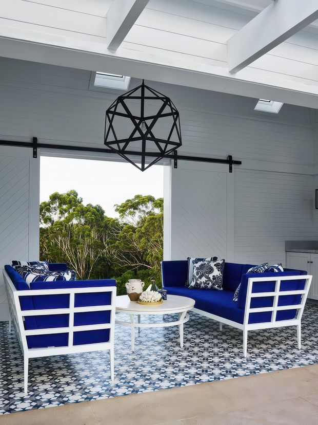 Bright, airy living room with white shiplap walls and exposed white ceiling beams, large sliding barn-style doors, and a geometric black pendant light. A vivid cobalt-blue and white sofa arrangement surrounds a round white coffee table on a patterned blue-and-white rug. Large open glazing frames leafy trees outside, while skylights add natural light.