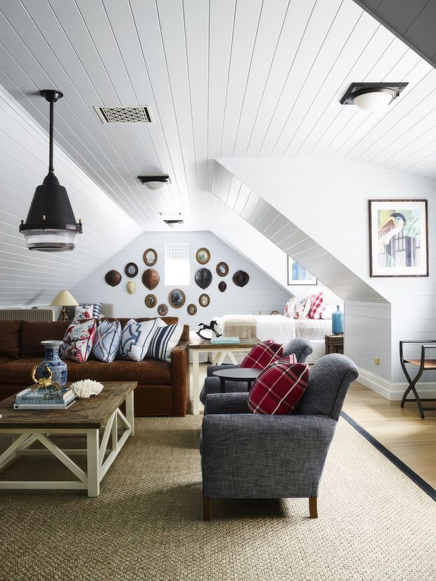 Bright attic-style living room with a sloped white beadboard ceiling and matching white walls, exposed roof geometry, and a small dormer-like window. The room contains a brown leather sofa, patterned cushions, two charcoal upholstered armchairs with red plaid pillows, a rustic wood-and-painted coffee table, and a light carpet. Decorative plates and framed artwork add character, while a mix of pendant and flush-mounted ceiling lights provides illumination.