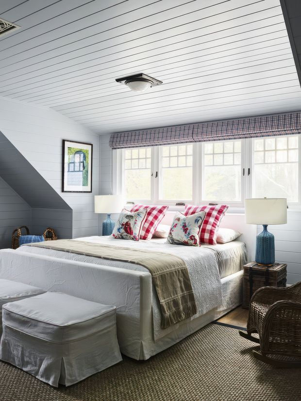 Bright coastal-style bedroom with white painted horizontal wall and ceiling panelling, a pitched or sloping ceiling area, and a bank of multi-pane windows dressed with a patterned Roman blind. The room contains a white upholstered bed layered with red-and-white checked and floral cushions, matching bedside lamps with blue ceramic bases, woven wicker seating, a fabric-covered bench, and a textured neutral rug.