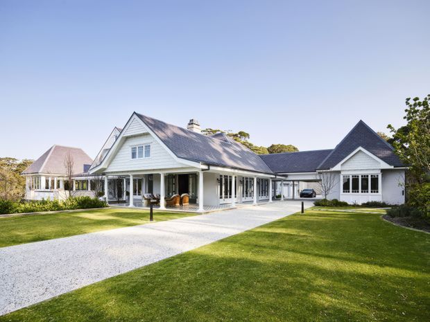A large, single-storey residential house photographed from the front, set within expansive manicured lawns. The home features white weatherboard cladding, multiple steep gabled roof forms with dark grey roofing, covered verandas, tall traditional windows, and a broad pale gravel driveway leading to a garage or carport wing. The architecture has a refined traditional, coastal-style character.
