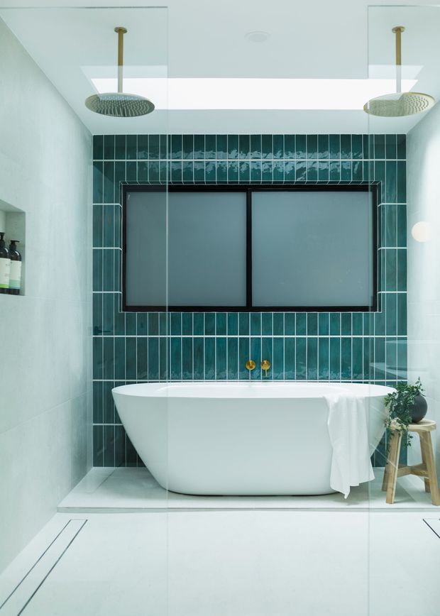 A refined contemporary bathroom featuring a freestanding white bathtub, dark teal vertically arranged wall tiles with contrasting grout, a large frosted window, twin rainfall showerheads reflected in glass, brass wall-mounted tap fittings, pale tiled flooring, and a small timber stool with greenery. The space is bright, clean, and spa-like.