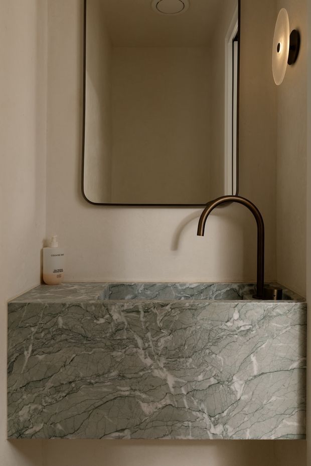 Close-up view of a compact bathroom vanity featuring a deep rectangular basin clad in dramatic grey-green veined natural stone. A dark bronze curved faucet, frameless rounded-corner mirror, recessed ceiling vent, and minimalist wall sconce sit against warm beige plaster walls. The styling is restrained, refined, and softly lit.