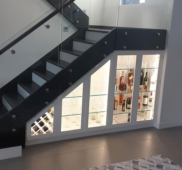 Interior view of a contemporary stairway with dark painted stringers and treads, clear glass balustrade panels, and integrated illuminated glass-front cabinetry beneath the stairs displaying wine and spirits. The space has pale walls, a smooth grey floor, and a modern built-in bar or wine-storage feature.