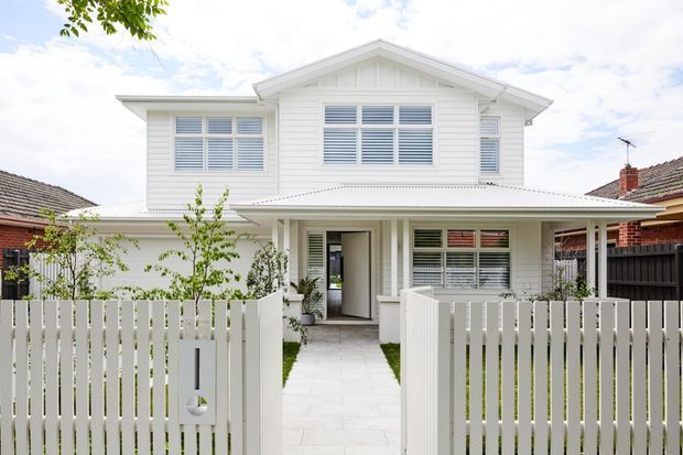 A bright white two-storey residential home viewed from the street, featuring weatherboard cladding, a prominent front gable, covered entry porch, louvred and shuttered windows, and a white picket fence with a paved path leading to the entrance. Neighbouring brick homes are visible at either side.