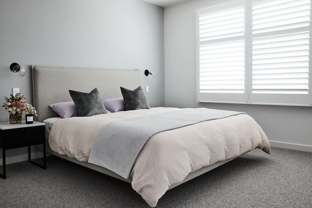 A bright, calm bedroom with a low upholstered bed dressed in pale neutral bedding, layered lavender and charcoal cushions, and a dark bedside table with a floral arrangement. Soft grey walls, grey carpet, wall-mounted reading lights, and large white plantation shutters create a clean contemporary interior.