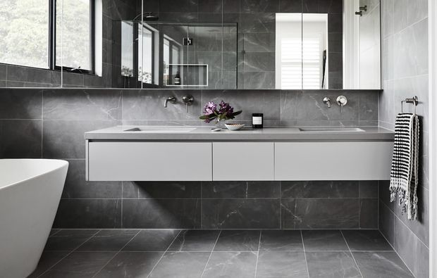 Contemporary bathroom with a wide floating double vanity, white integrated basins, dark grey marble-look wall and floor tiles, and a large mirrored wall. A freestanding white bathtub is partially visible at left, with a window and white shutter providing natural light.