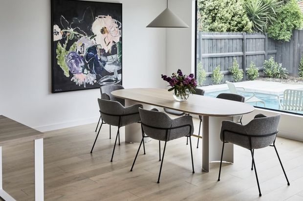 A bright contemporary dining area with a pale timber dining table and sculptural pedestal base, surrounded by upholstered charcoal dining chairs. A large abstract artwork and minimalist pendant light add visual interest, while full-height glazing opens views to a fenced outdoor patio and swimming pool.