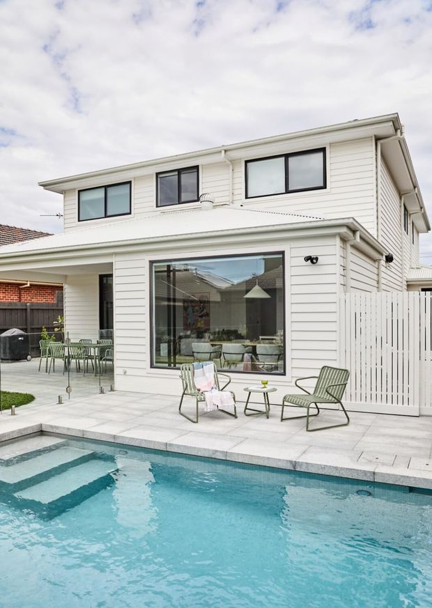 Exterior view of a two-storey white weatherboard house overlooking a swimming pool. The home has a shallow pitched roof with eaves, large black-framed windows and glazed doors, a tiled pool terrace, white privacy fencing, and outdoor lounge and dining furniture. A bright, contemporary suburban setting is visible.