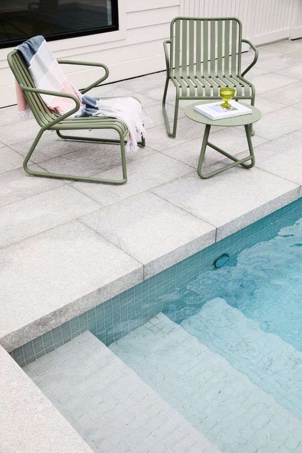 A contemporary poolside terrace with pale square paving surrounding a turquoise tiled swimming pool. Two sage-green metal lounge chairs and a matching side table sit beside the pool, with a folded striped throw on one chair and a book and glass on the table. White horizontal wall cladding and a dark-framed window form the backdrop.