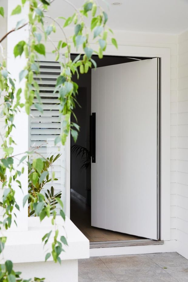 A bright residential entry viewed from outside, featuring a large white pivot front door with a black handle set into a white horizontal-clad facade. The recessed doorway opens onto a dark interior hallway, with white louvre blinds beside the entrance, pale tiled paving, and leafy plants in the foreground.