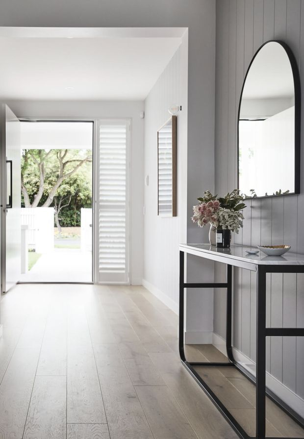 Bright, refined entry hall with pale timber flooring, white walls and plantation shutters. A slim black console table sits against a grey vertical-panelled wall beneath a large arched black-framed mirror, with a floral arrangement providing a soft natural accent. Glazing and an open doorway connect the hall to a bright garden-facing exterior.