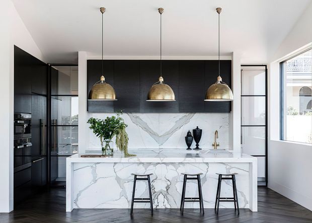 A refined contemporary kitchen featuring a large marble-look island with seating for three, dark full-height cabinetry, a matching dramatic stone splashback, and three brass pendant lights. Black-framed glazing and dark timber flooring add contrast to the bright white surfaces.